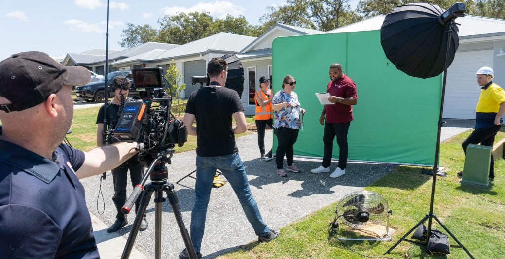 The Video Box team on set for a recent campaign for Catapult Homes: A camera operator holds a video camera, while a sound producer and stills camera photographer wait for instruction. The director works with actor Sam Thaiday on the video script in front of a green screen, held up by two production assistants. Video equipment such as lighting, stands, a fan and other cables can be seen, while homes build by Catapult are featured in the background.