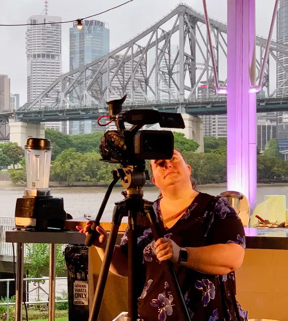 The Video Box director, Sarah Robinson, captures video production content for a cooking demonstration held in Brisbane, Australia by the Story Bridge using a video camera. The Story Bridge is seen in the background, including city.