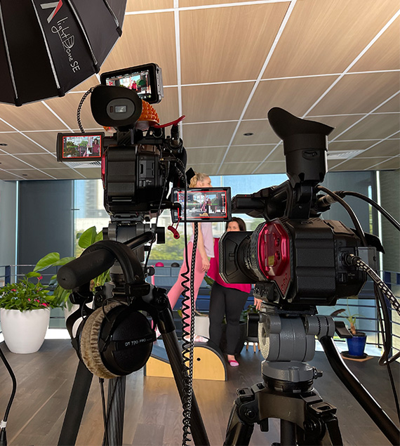 Two Panasonic DVX 200 Video Cameras on tripods are focused on a Pilates teacher instructing a studio on a High Barrel Arc. A Lighting Soft Box is seen in the left top corner, lighting the scene being recorded by the Video Cameras. The cameras have their monitors out giving the audience a glimpse into which camera is recording what video footage.