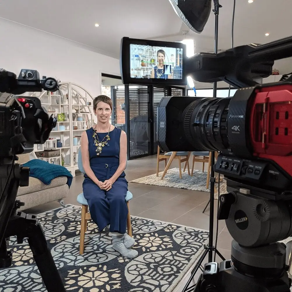 Helen Joy Butler sitting behind two red Panasonic video cameras with video lights in her lounge room during a video production session.