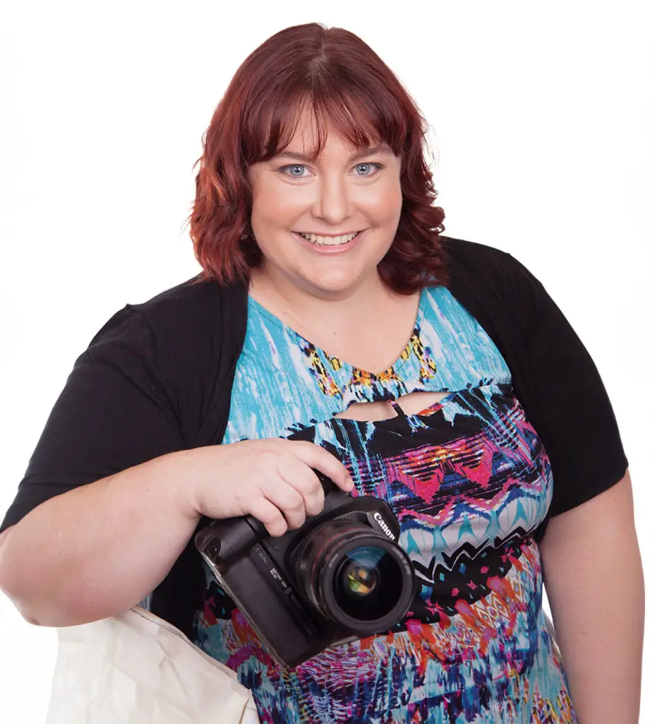 Sarah Robinson sitting in a white chair, wearing a blue top and black jacket, holding a Canon camera.