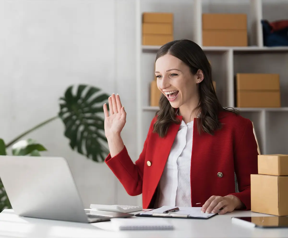 A brunette woman sitting in an office, smiling and waving during a Zoom call, creating a welcoming and engaging atmosphere.