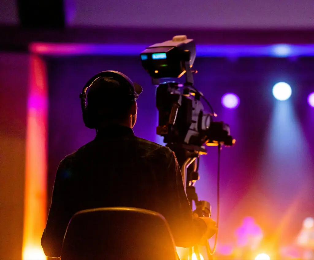 A camera operator sitting and holding the panning arm of a camera tripod with a professional video camera aimed at a stage, with blurred stage lighting in the background.