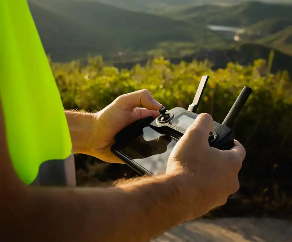 Close-up of a licensed drone operator’s hands holding a drone remote control with antennas up, wearing a safety jacket and standing on a hill with scenic bushes and a rolling landscape in the background.