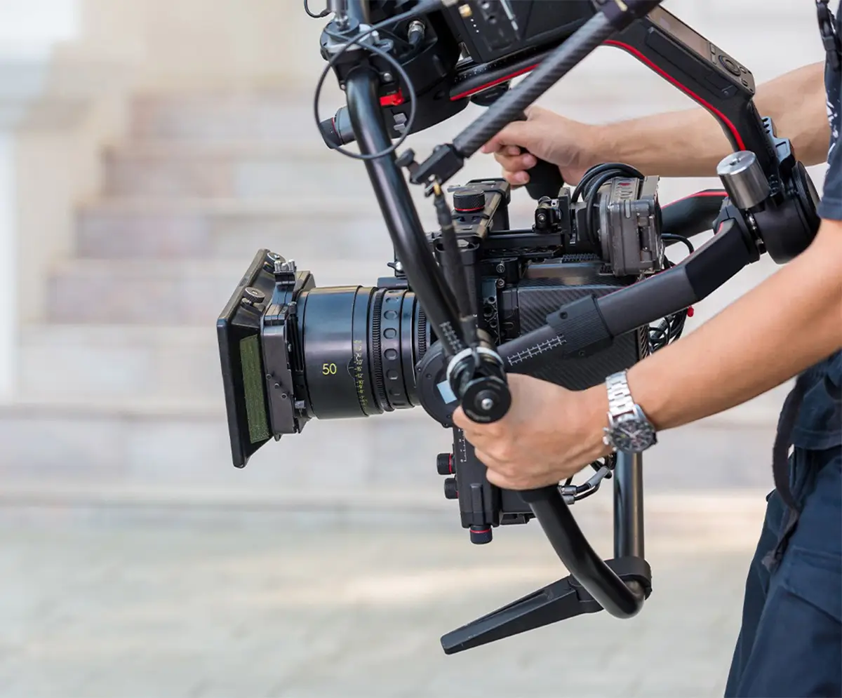 Modern camera equipment on a carbon fiber DJI Ronin frame with a professional 3-axis stabilization system, operated by a camera technician’s hands during a panning shot.