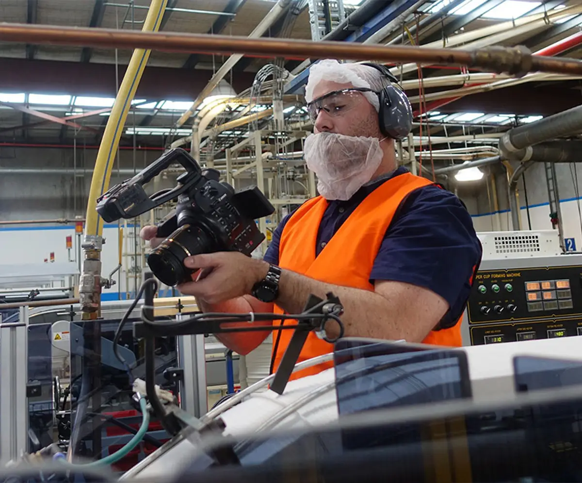 A camera operator filming at a paper cup manufacturing plant, wearing a mask and safety gear while holding a professional video camera for a panning shot.