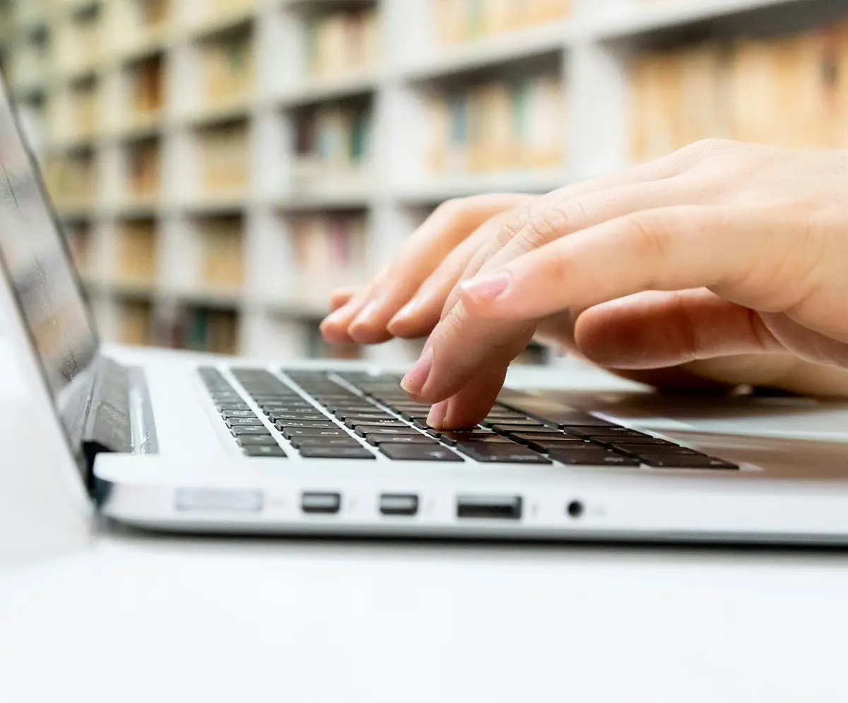 Close-up of hands typing on a laptop keyboard, set on a white desk with a corporate bookcase in the background.