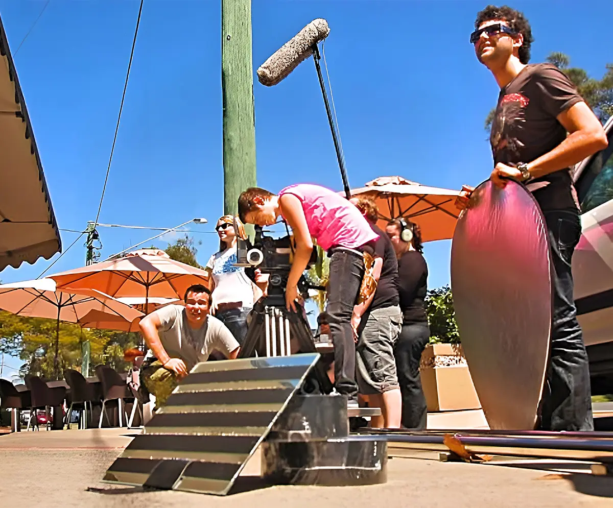 The Video Box production team filming at a local cafe, featuring a man holding a reflector, a sound operator with a boom pole, and the director of photography adjusting the camera on a dolly track.