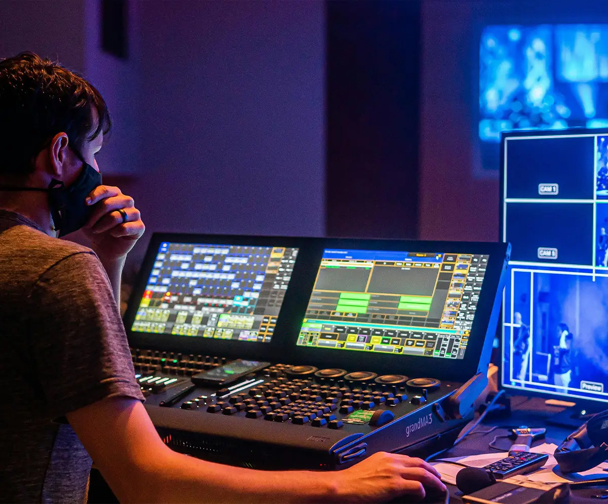 A masked post-production technician operating a live production desk with multiple buttons and screens, watching a large preview monitor while managing the technical elements.