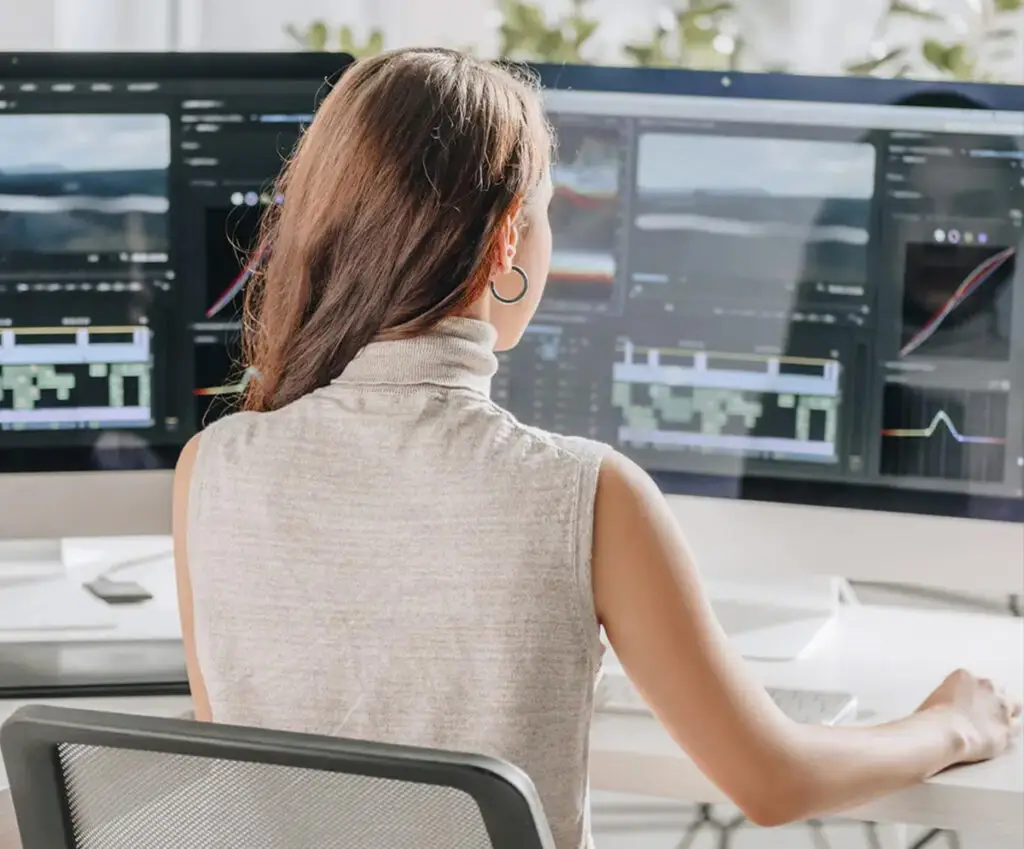 A woman sitting at a bright and airy post-production desk with multiple Apple monitors displaying Premiere Pro editing software, using a mouse to navigate the screens.