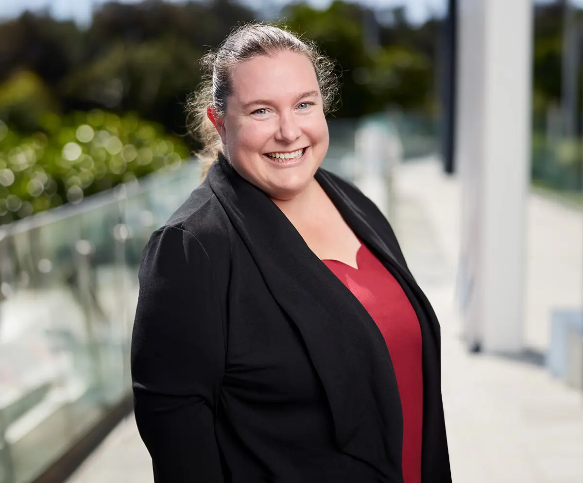 Headshot of Sarah Robinson, Company Director of The Video Box, wearing a suit jacket and red shirt, smiling outdoors with green trees and a corporate office setting blurred in the background.