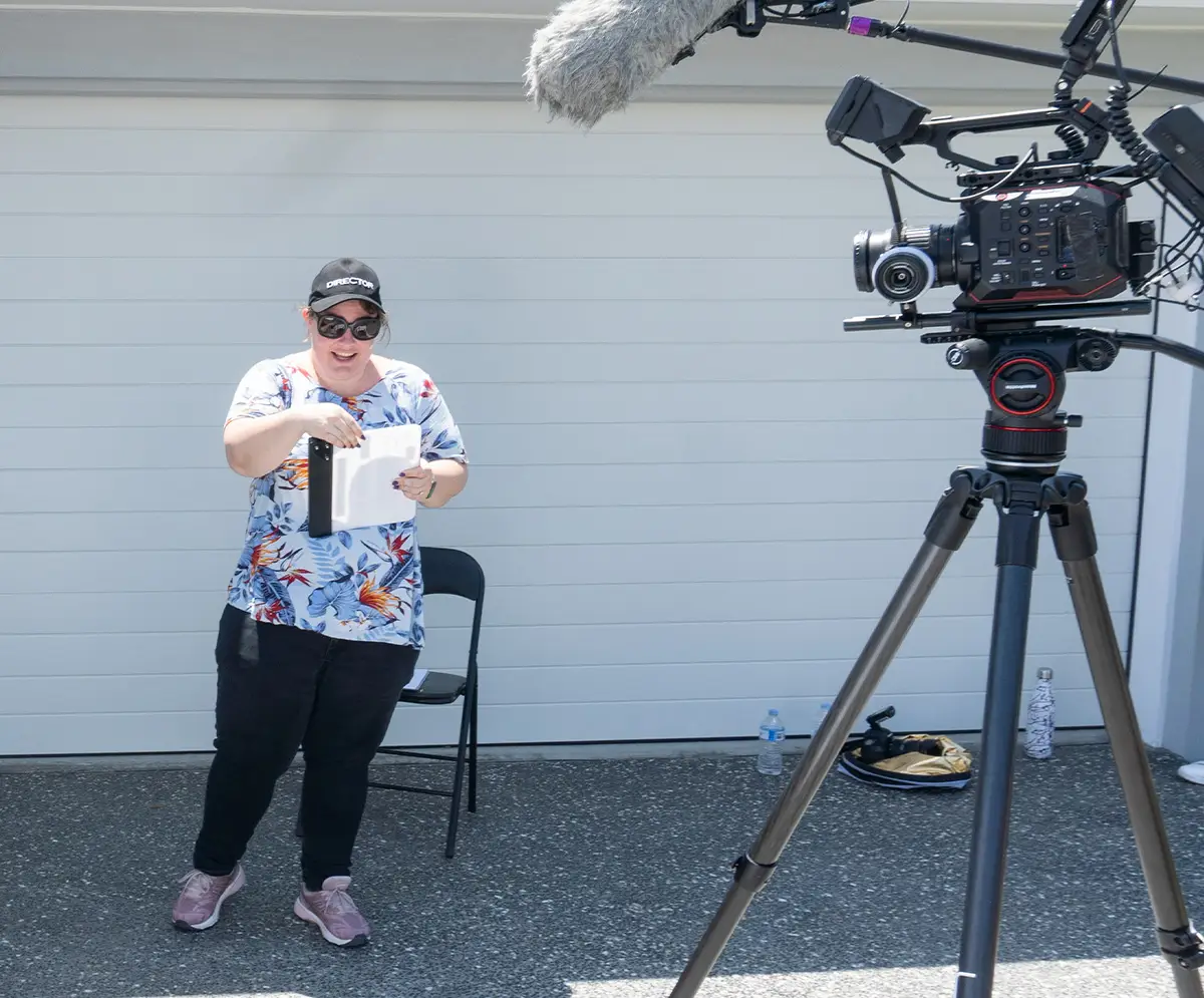 Sarah Robinson, wearing a director hat and holding a clapperboard, standing beside a professional video camera and tripod. A garage with a chair and video production equipment is visible in the background.