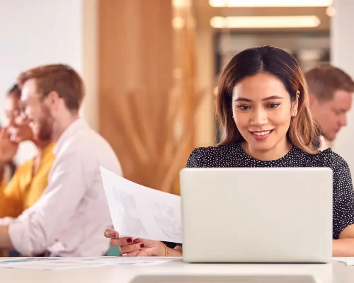 A woman smiling at her computer while holding a document at her desk, with coworkers visible in the background.