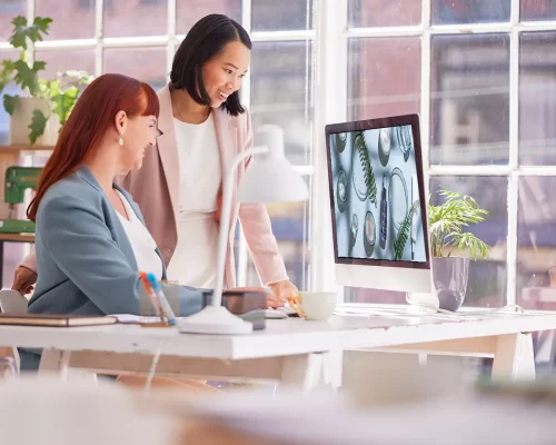 Two female marketers smiling as they watch a beauty product video on an Apple monitor in a light and airy office with large windows. The desk is white with a lamp, pot plant, and general office supplies.