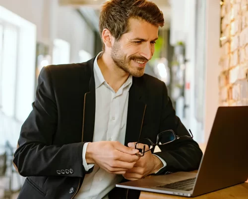 A man in a business suit casually leaning against his desk, smiling at his laptop while holding his glasses in one hand. His modern corporate office with open windows and a brick wall is blurred in the background.