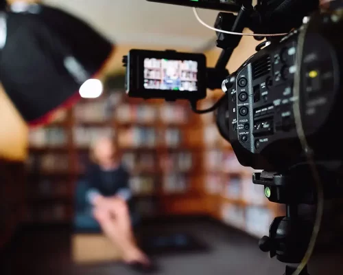 A professional video camera recording a subject seated in front of a bookcase, with a blurred Aperture light overhead.