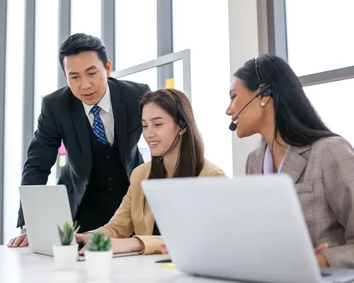 A manager and employee smiling at their desk while reviewing successful results on a computer screen, with a coworker looking on.